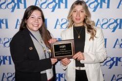 Two college students dressed professionally standing in front of a white backdrop with blue lettering. They are holding an award.